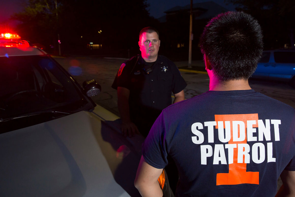 University of Illinois police officer talking with Student Patrol member alongside squad vehicle with lights activated at night.